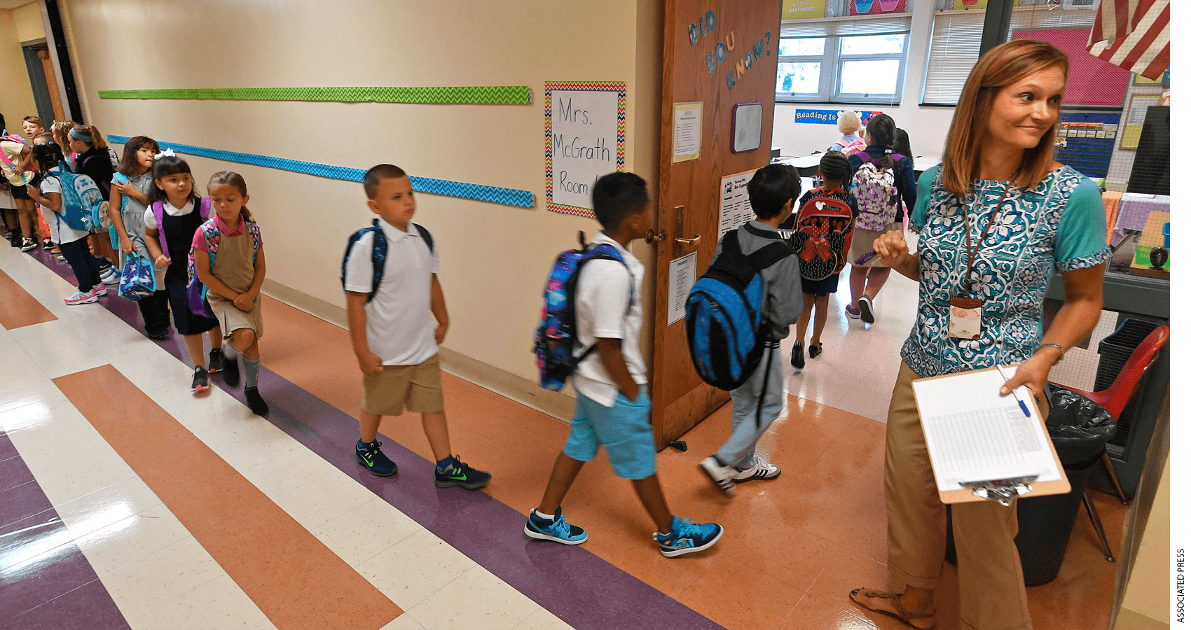 Teacher holding clipboard welcomes students as they enter classroom