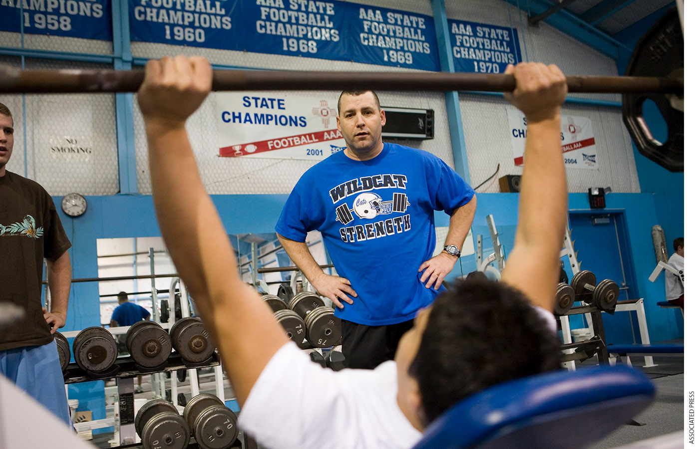 A high-school football player lifts weights while his coach watches.