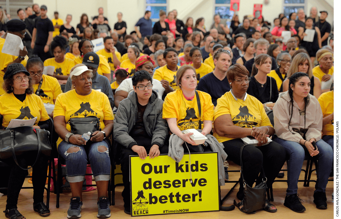 In 2019, parents opposed to a proposed merger of several elementary schools in Oakland, California, attended an Oakland Unified School District board meeting. Attendees in yellow shirts, as well as the one holding a sign, were members of The Oakland REACH.