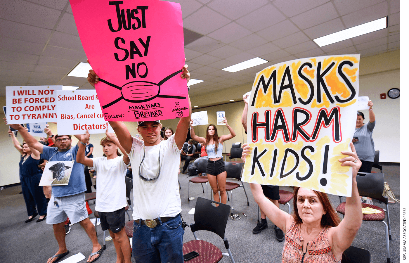 Room full of people holding signs that say "Masks Harm Kids", "Just Say No: Follow Mask Wars Brevard Instagram", "School Board Members Are Bias, Cancel culture Dictators!" and "I will not be forced to comply to tyranny."
