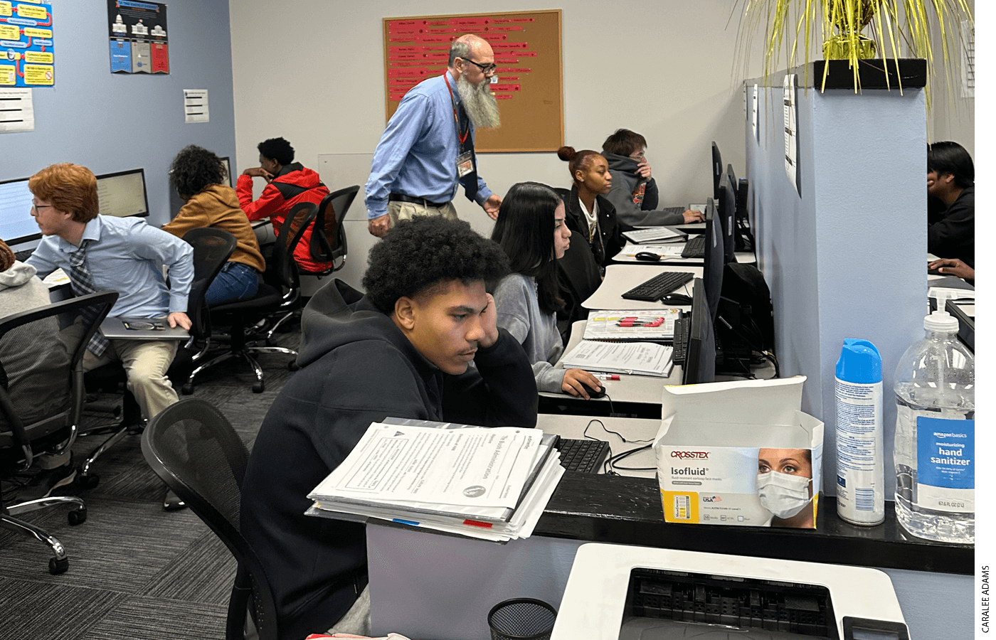 Social studies teacher Robert Carr (standing) checks students’ progress at computer workstations, while tutor John Brandon works with a teen one-on-one.