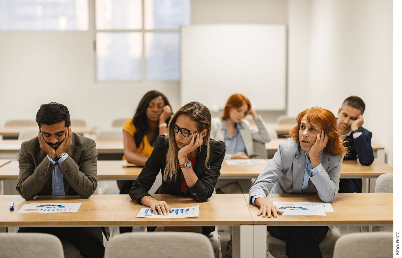 Six teachers sitting in two rows at desks in a professional development classroom looking bored and sleepy
